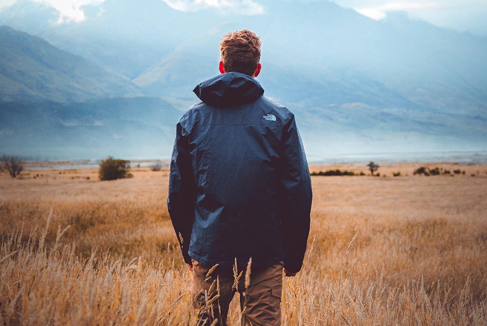 man, wearing a coat, stood in a field looking at a view of mountains